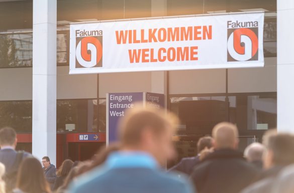 Crowd in front of a building with a large 'WILLKOMMEN WELCOME' banner and signs for 'Entrance West' (Fakuma event).